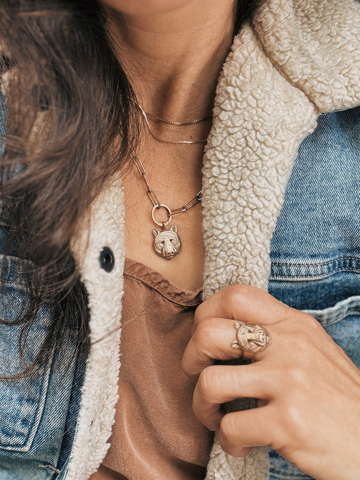 Close-up of a person wearing a necklace with a bear pendant and a ring, with a denim jacket and beige fleece lining.