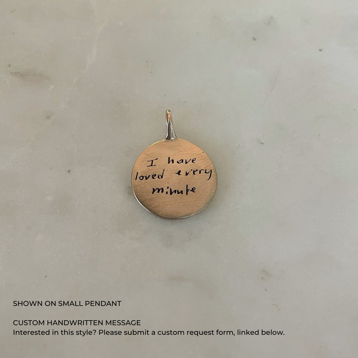 A small, round, bronze pendant necklace featuring a custom handwritten message. &