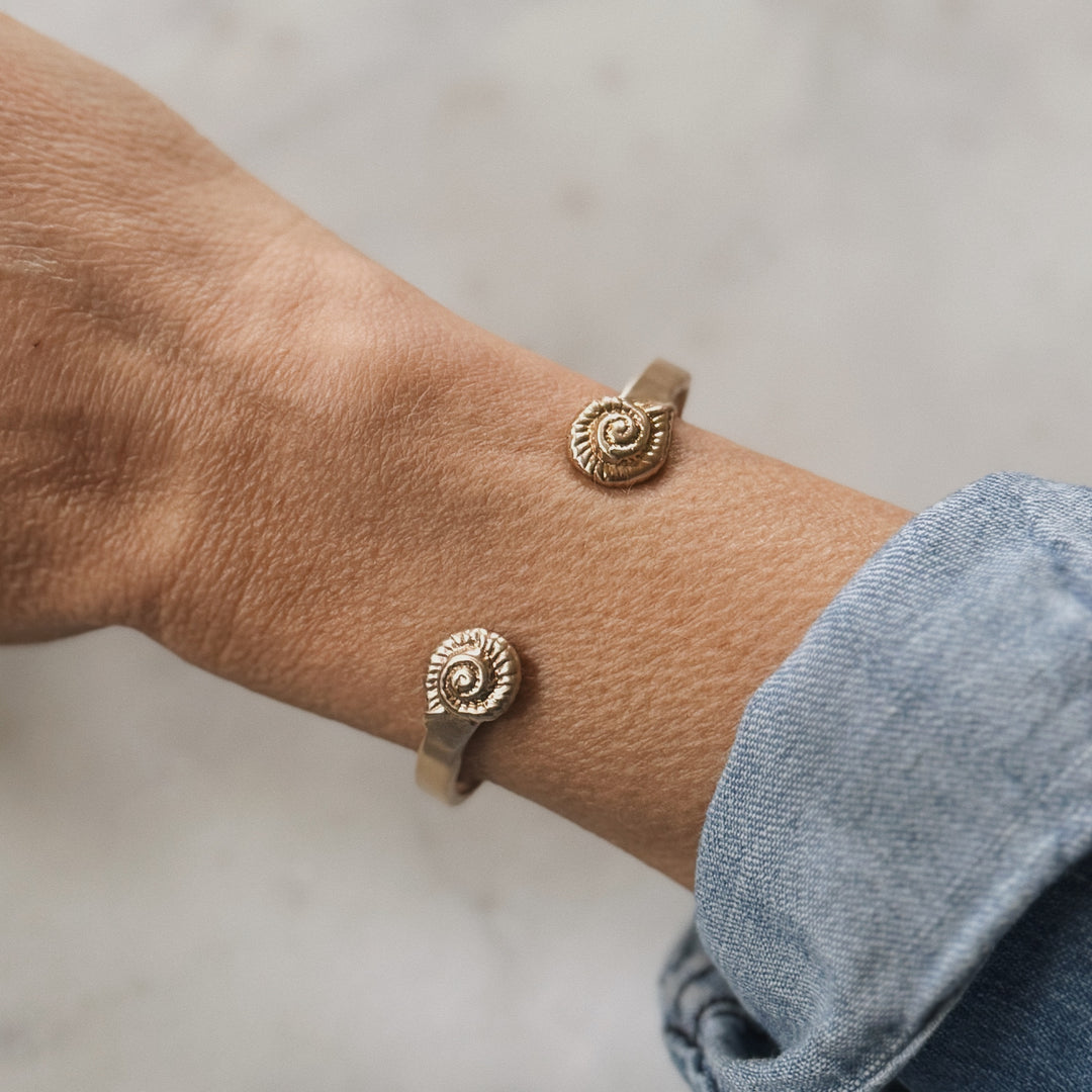 Close-up of a wrist wearing two bronze spiral-shaped snail shells on a neutral background