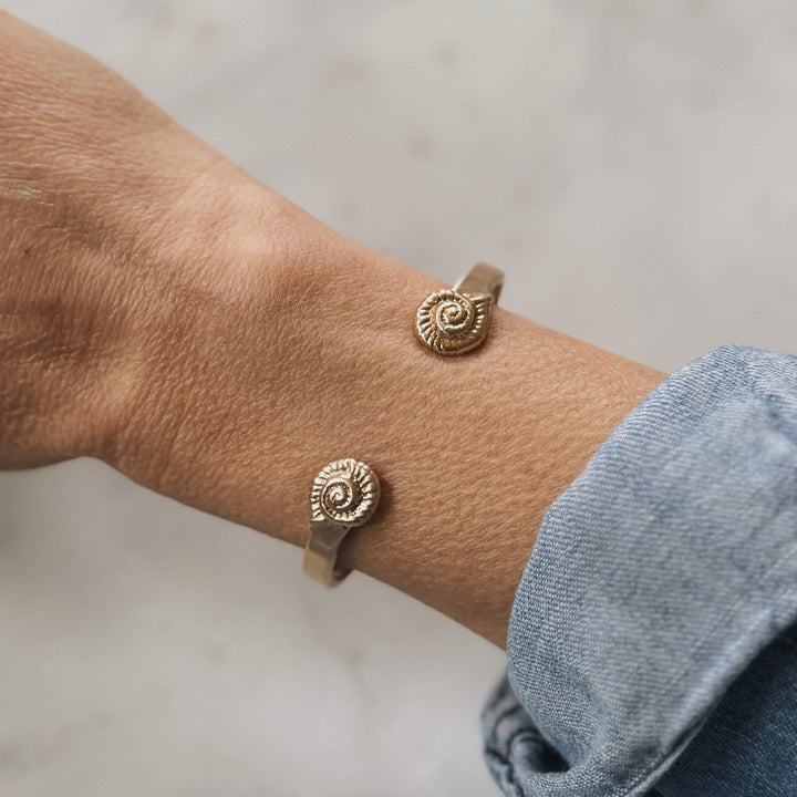 Close-up of a wrist wearing two bronze spiral-shaped snail shells on a neutral background
